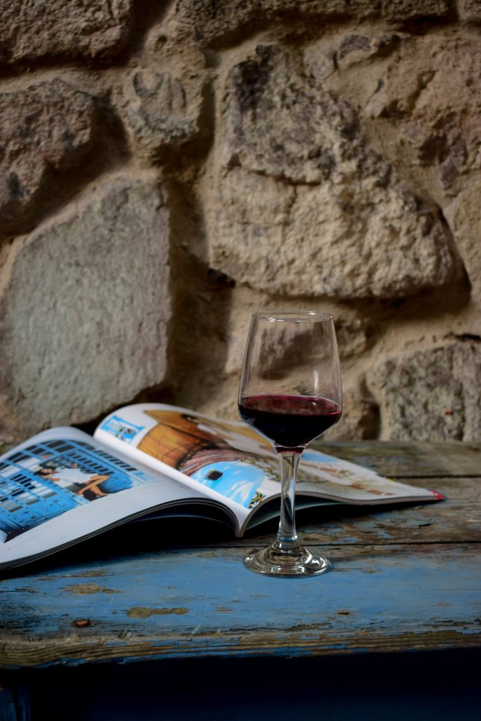 A glass of red wine on a wooden table beside an open travel magazine against a stone wall.