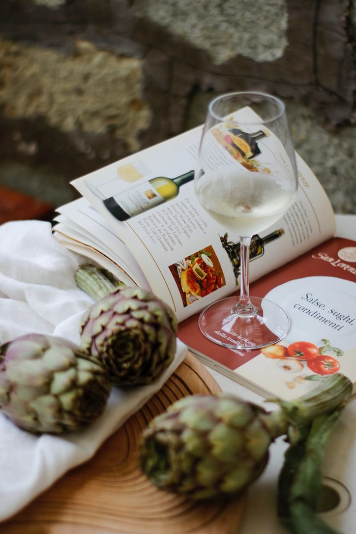 A glass of white wine with artichokes and magazine on rustic table setting.